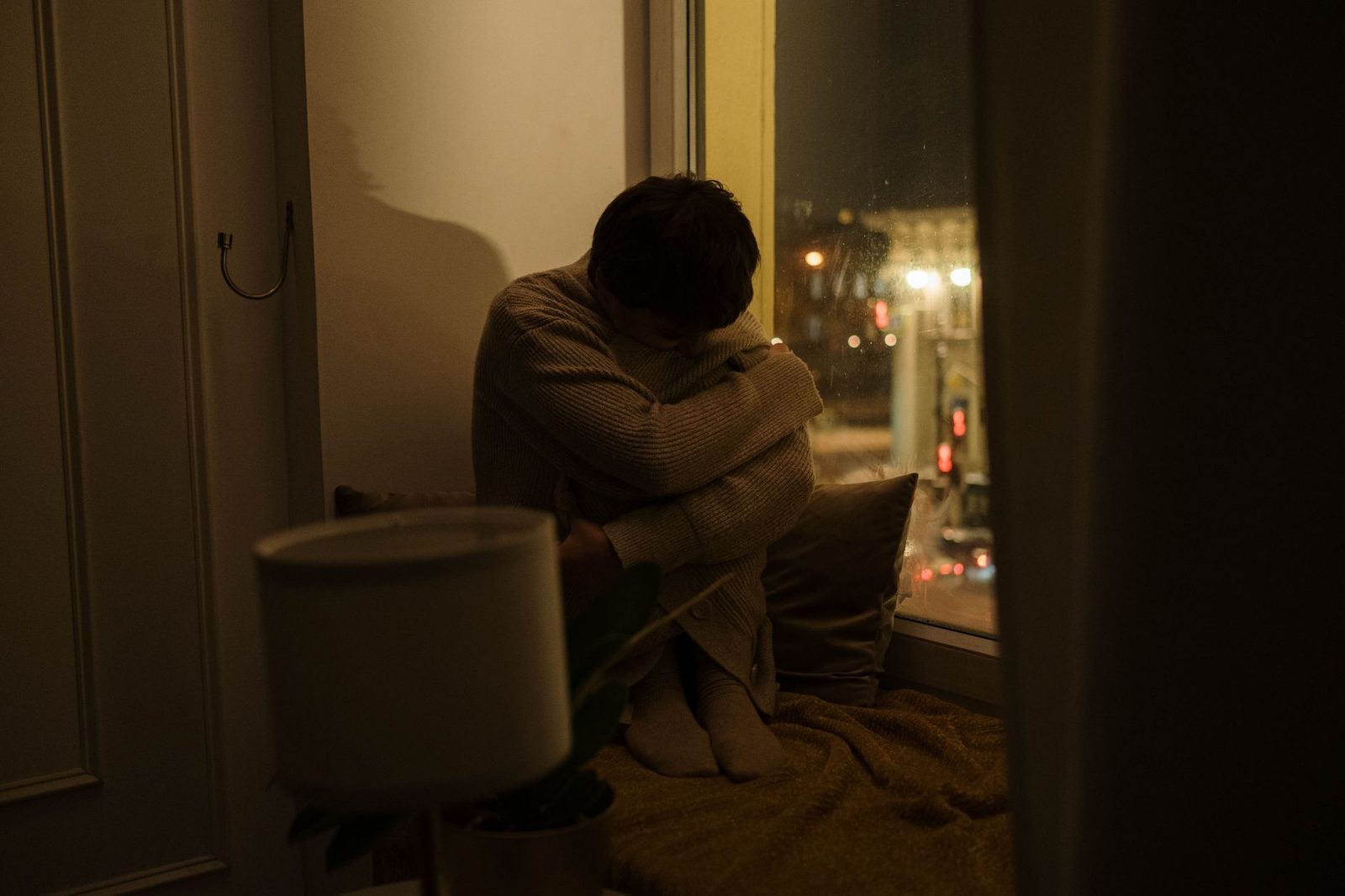 Gece yarısı pencere kenarında oturan bir kişinin uzaktan çekilmiş silueti — yalnızken ağırlaşan iç sıkıntısını anlatan sakin sahne
